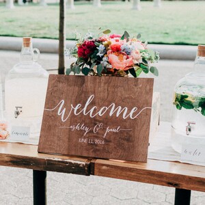 May include: A wooden sign with the text "Welcome Ashley & Paul June 11, 2018" on a table with a floral arrangement, two glass beverage dispensers, and a second wooden sign with a wedding ceremony schedule.