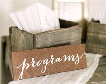 May include: A wooden sign with the word "programs" written in white calligraphy on a brown background. The sign is sitting on a white cloth and a wooden table. There is a wooden box filled with white paper programs behind the sign.