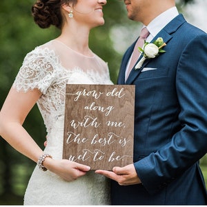 May include: A wooden sign with the words "grow old along with me, the best is yet to be" in white script. The sign is held by a couple, the bride in a white lace dress and the groom in a navy blue suit.