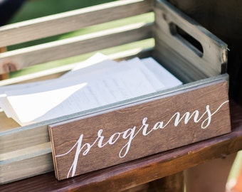 May include: A wooden crate filled with white paper programs for a wedding ceremony. The crate is sitting on a wooden table with a sign that says "programs" in white cursive lettering.