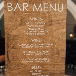 May include: A wooden sign with a white bar menu listing alcoholic beverages. The sign reads "BAR MENU" at the top and lists spirits, wine, and beer options.