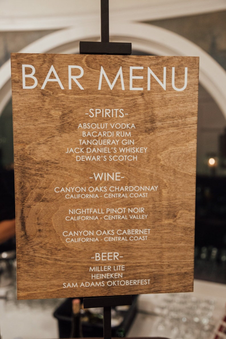 May include: A wooden sign with a white bar menu listing alcoholic beverages. The sign reads "BAR MENU" at the top and lists spirits, wine, and beer options.