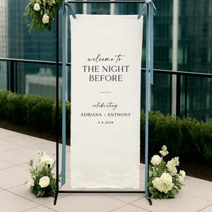 May include: A wedding welcome sign with the text "welcome to THE NIGHT BEFORE" in elegant script. The sign is white, framed in black, and adorned with blue ribbons and floral arrangements. The names Adriana + Anthony and the date 3.4.2024 are also displayed.