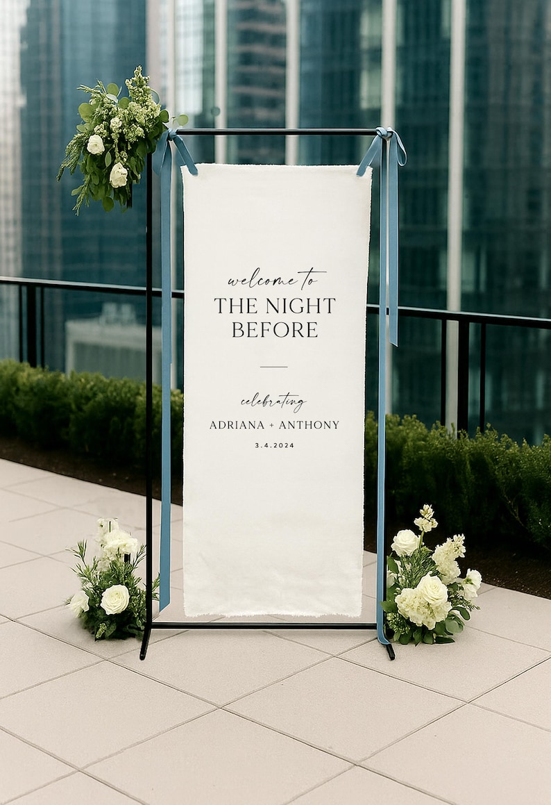 May include: A wedding welcome sign with the text "welcome to THE NIGHT BEFORE" in elegant script. The sign is white, framed in black, and adorned with blue ribbons and floral arrangements. The names Adriana + Anthony and the date 3.4.2024 are also displayed.