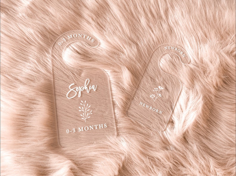 Pu&ograve; includere: Due appendiabiti in acrilico bianco con un design floreale. Un appendiabiti &egrave; etichettato "Neonato" e l'altro "Sophia 0-3 mesi".