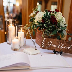 Peut inclure: Un livre d'or blanc avec une pancarte en bois sur laquelle est inscrit "Guestbook" en calligraphie blanche. La pancarte est d&eacute;cor&eacute;e de petites branches de fleurs blanches. Le livre est pos&eacute; sur une table blanche avec trois bougies dans des bougeoirs en verre.