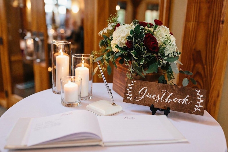 Peut inclure: Un livre d'or blanc avec une pancarte en bois sur laquelle est inscrit "Guestbook" en calligraphie blanche. La pancarte est d&eacute;cor&eacute;e de petites branches de fleurs blanches. Le livre est pos&eacute; sur une table blanche avec trois bougies dans des bougeoirs en verre.