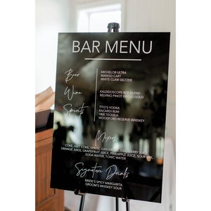 May include: Black acrylic sign with white text listing a bar menu. The menu includes beer, wine, spirits, mixers, and signature drinks. The signature drinks are "Bride's Spicy Margarita" and "Groom's Whiskey Sour".