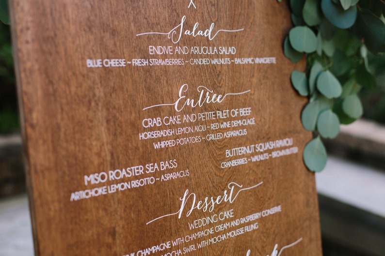 May include: A wooden sign with a white handwritten menu for a wedding reception. The menu includes salad, entree, and dessert options. The sign is on a wooden stand and has a rustic look.