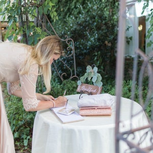 Peut inclure: Une femme en robe rose p&acirc;le signe un livre d'or blanc sur une table blanche avec une pancarte en bois qui dit "Livre d'or" et une pochette rose.