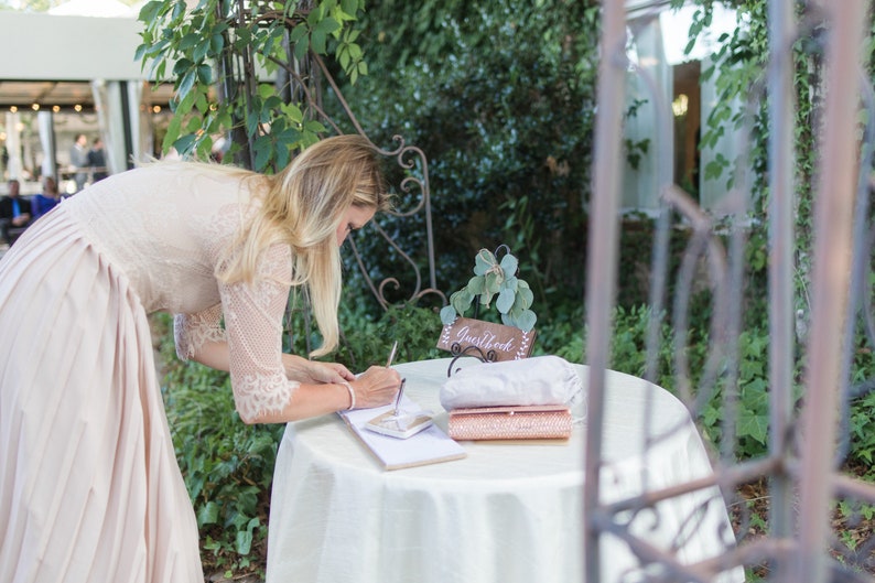 Peut inclure: Une femme en robe rose p&acirc;le signe un livre d'or blanc sur une table blanche avec une pancarte en bois qui dit "Livre d'or" et une pochette rose.