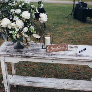 Peut inclure: Une table en bois blanc avec un arrangement floral blanc, une lanterne avec une bougie, une pancarte indiquant "Guestbook" et un livre d'or blanc avec un stylo sur le dessus.
