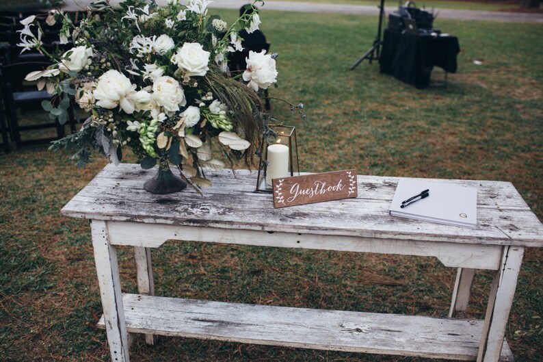 Peut inclure: Une table en bois blanc avec un arrangement floral blanc, une lanterne avec une bougie, une pancarte indiquant "Guestbook" et un livre d'or blanc avec un stylo sur le dessus.