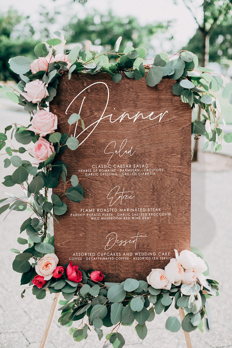 May include: A wooden sign with a white calligraphy script that reads "Dinner" and lists the menu for a wedding reception. The menu includes salad, entree, and dessert options. The sign is decorated with pink and white flowers and greenery.