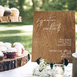 May include: A wooden sign with the text "signature cocktails" written in white cursive. The sign lists two cocktails: Paloma and Moscow Mule, with their ingredients.