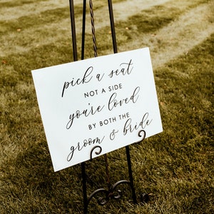 Puede incluir: Un cartel blanco con texto en caligraf&iacute;a negra sobre un caballete en un campo de hierba. El cartel dice "pick a seat not a side you're loved by both the groom & bride".