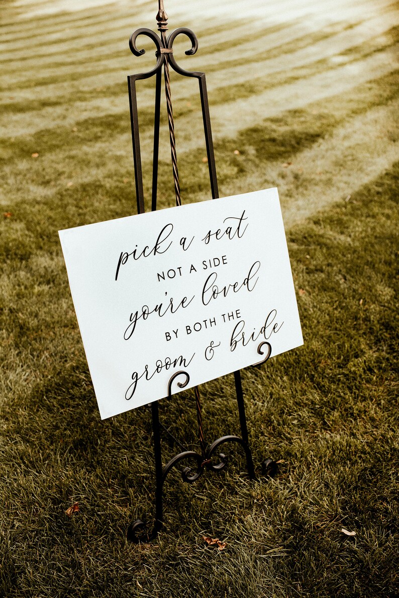 Puede incluir: Un cartel blanco con texto en caligraf&iacute;a negra sobre un caballete en un campo de hierba. El cartel dice "pick a seat not a side you're loved by both the groom & bride".