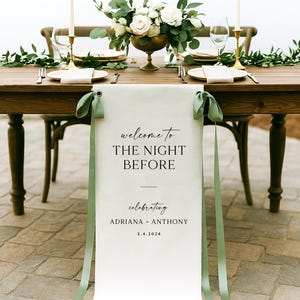 May include: A beautifully set table for a wedding celebration. The wooden table is adorned with a floral centerpiece, candles, and place settings. A banner reads "welcome to THE NIGHT BEFORE" celebrating Adriana + Anthony, 3.4.2024.