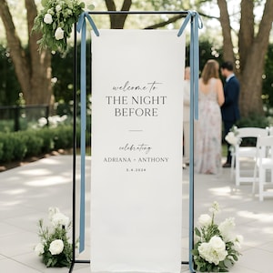 May include: A white wedding welcome sign with black text that reads "welcome to THE NIGHT BEFORE celebrating ADRIANA + ANTHONY 3.4.2024". The sign is on a black metal frame with blue ribbons and floral arrangements.
