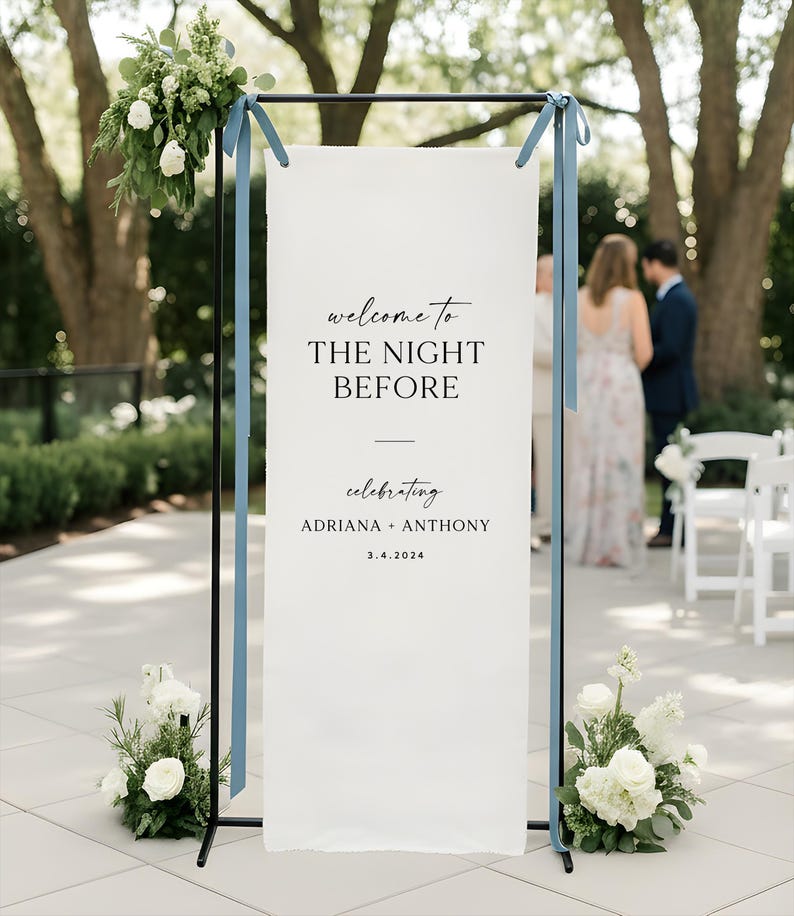 May include: A white wedding welcome sign with black text that reads "welcome to THE NIGHT BEFORE celebrating ADRIANA + ANTHONY 3.4.2024". The sign is on a black metal frame with blue ribbons and floral arrangements.