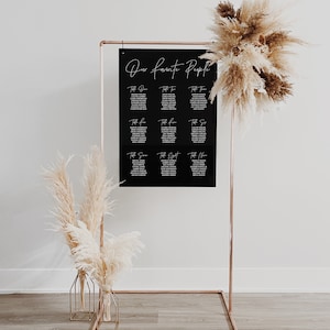 May include: A black acrylic seating chart with white calligraphy that reads "Our Favorite People" and lists table numbers and guest names. The chart is displayed on a copper stand with two dried pampas grass arrangements in gold vases.