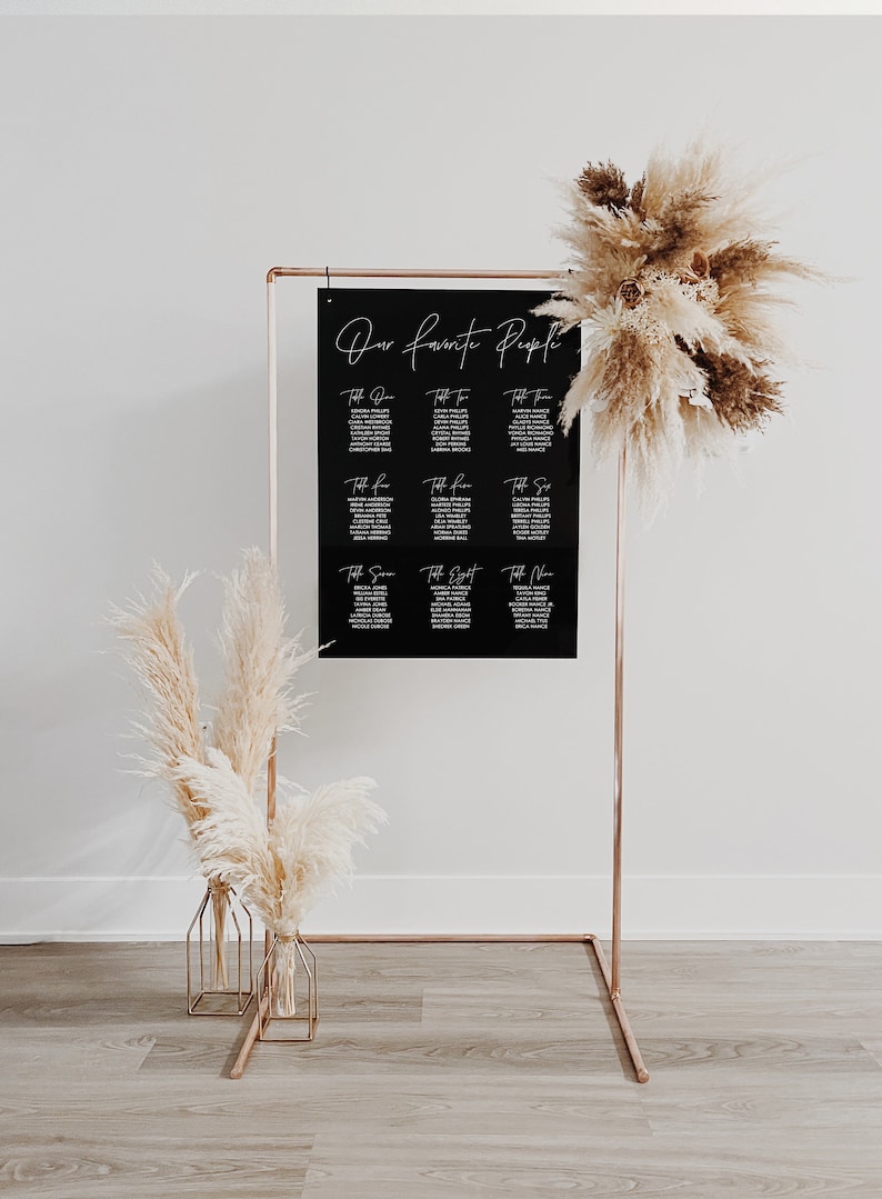 May include: A black acrylic seating chart with white calligraphy that reads "Our Favorite People" and lists table numbers and guest names. The chart is displayed on a copper stand with two dried pampas grass arrangements in gold vases.