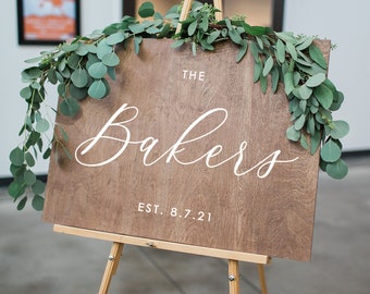 May include: A wooden sign with the text "The Bakers Est. 8.7.21" in white lettering. The sign is decorated with greenery and is displayed on a wooden easel.