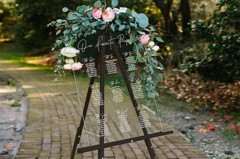 May include: A clear acrylic wedding seating chart with the text "Our Favorite People" at the top. The chart is on a black easel and decorated with greenery and pink flowers.