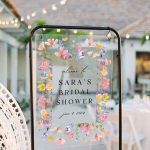 May include: A clear acrylic sign with a floral border and the text "Welcome to Sara's Bridal Shower June 8, 2024".