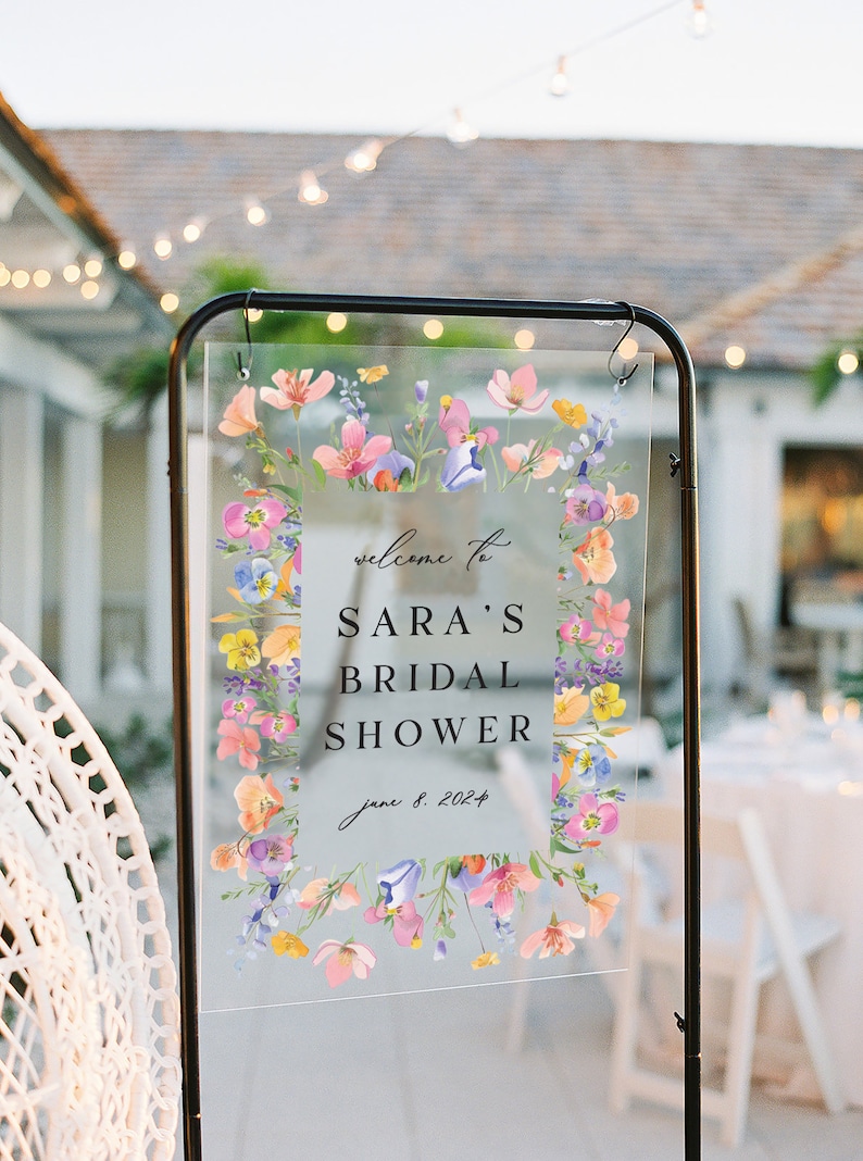 May include: A clear acrylic sign with a floral border and the text "Welcome to Sara's Bridal Shower June 8, 2024".