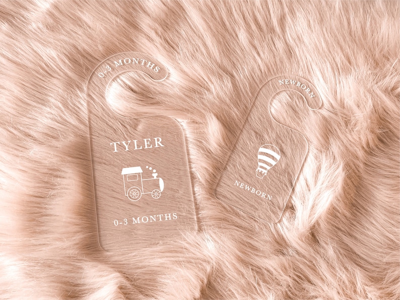 May include: Two clear acrylic baby clothes hanger tags with white text and graphics. One tag has a train graphic and the text "TYLER" and "0-3 MONTHS". The other tag has a hot air balloon graphic and the text "NEWBORN".