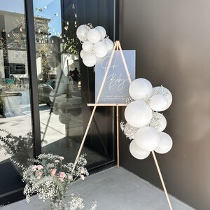 Peut inclure: Un chevalet en bois avec une pancarte blanche sur laquelle est inscrit "Oh Baby!" avec une date et un nom. Le chevalet est d&eacute;cor&eacute; de ballons blancs et de gypsophile.