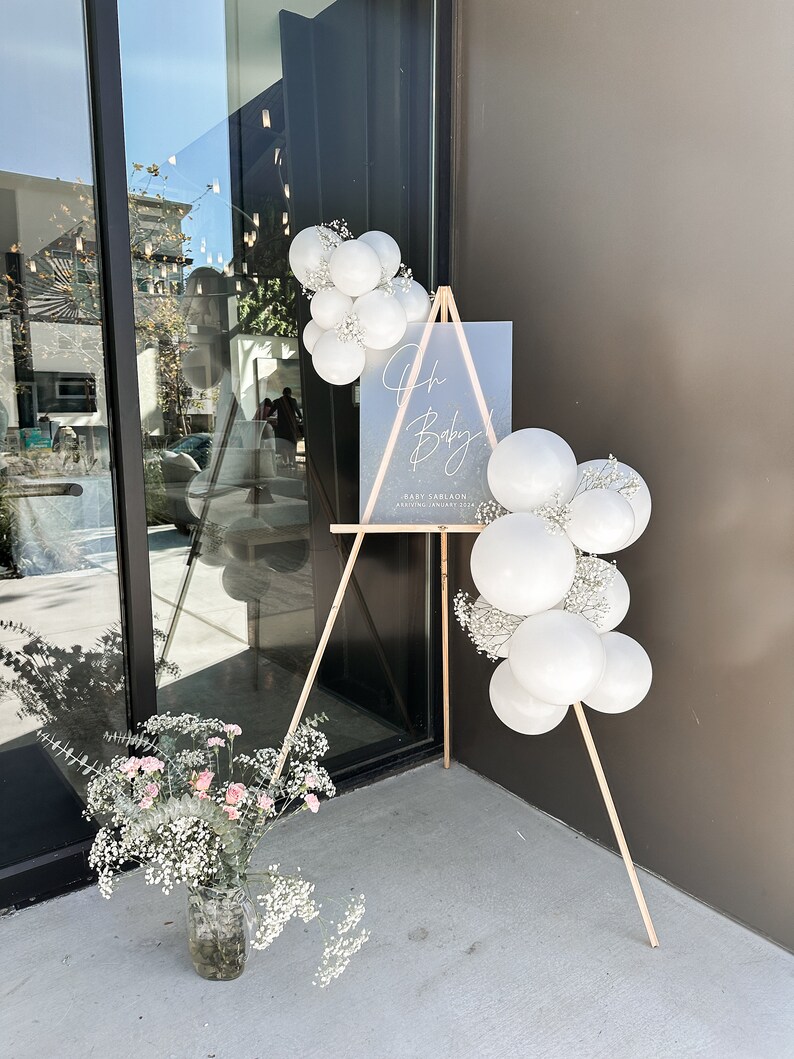 Peut inclure: Un chevalet en bois avec une pancarte blanche sur laquelle est inscrit "Oh Baby!" avec une date et un nom. Le chevalet est d&eacute;cor&eacute; de ballons blancs et de gypsophile.