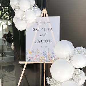 Puede incluir: Un cartel de bienvenida de boda con un fondo morado claro y un dise&ntilde;o floral en la parte inferior. El cartel dice "We're so happy you're here Sophia and Jacob 3.1.2024".