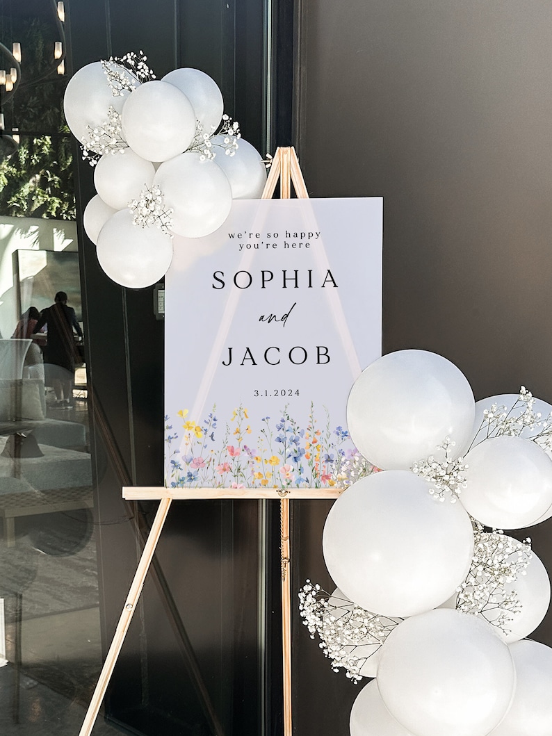 Puede incluir: Un cartel de bienvenida de boda con un fondo morado claro y un dise&ntilde;o floral en la parte inferior. El cartel dice "We're so happy you're here Sophia and Jacob 3.1.2024".