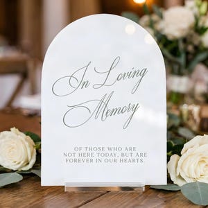 May include: An arched white acrylic sign with the text In Loving Memory of those who are not here today, but are forever in our hearts. The sign features elegant cursive and block lettering and is supported by a clear rectangular base.