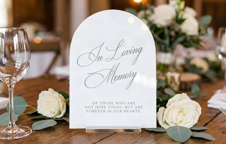 K&ouml;nnte beinhalten: Ein bogenf&ouml;rmiges wei&szlig;es Acrylschild mit dem Text In Loving Memory of those who are not here today, but are forever in our hearts. Das Schild zeigt eine elegante Schreibschrift sowie Blockschrift und steht auf einem klaren Sockel.