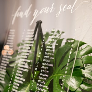May include: A clear acrylic seating chart with white lettering. The chart is titled "Find Your Seat" and lists names and table numbers. The chart is leaning against a green tropical plant.
