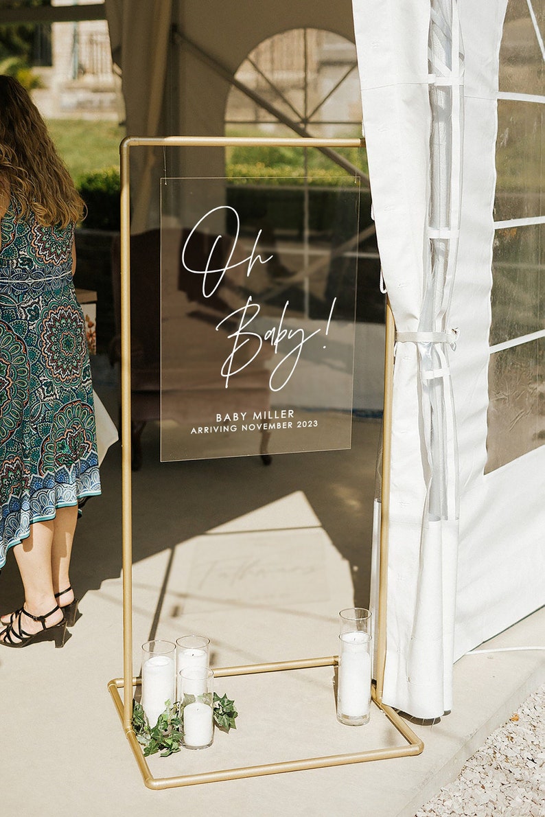 Peut inclure: Un support en m&eacute;tal dor&eacute; avec une pancarte en acrylique transparente qui indique "Oh Baby! Baby Miller Arriving November 2023". La pancarte est d&eacute;cor&eacute;e de calligraphie blanche. Le support est d&eacute;cor&eacute; de bougies blanches et de feuillage vert.
