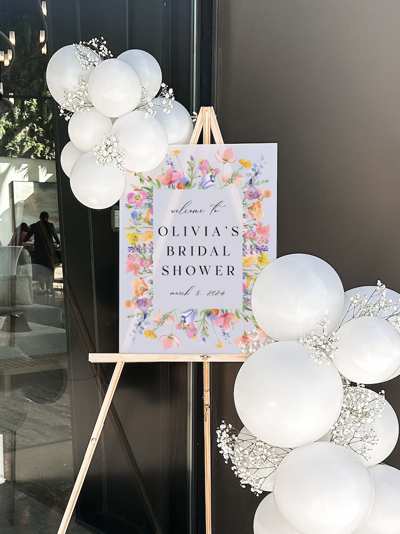 May include: A welcome sign for a bridal shower with a floral design and the text "Welcome to Olivia's Bridal Shower March 5, 2024". The sign is on an easel and surrounded by white balloons with baby's breath.