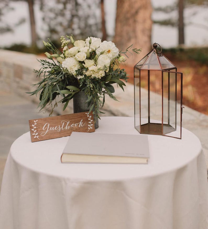 Peut inclure: Un livre d'or blanc avec une pancarte en bois indiquant "Guestbook" sur une nappe blanche avec un bouquet de fleurs blanches et une lanterne en verre.
