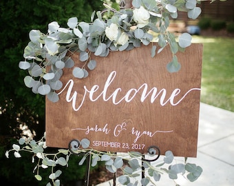May include: A wooden welcome sign with white calligraphy that reads "welcome", "sarah & ryan", and "SEPTEMBER 23, 2017". The sign is decorated with eucalyptus greenery and white flowers.