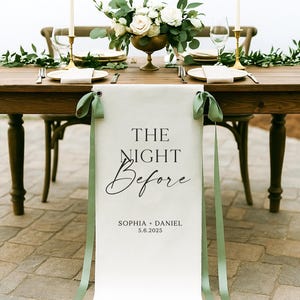 May include: A beautifully set wedding reception table with a floral centerpiece, candles, and place settings. A white banner with the text "THE NIGHT Before" and the names Sophia and Daniel, with the date 5.6.2025, hangs from the table.