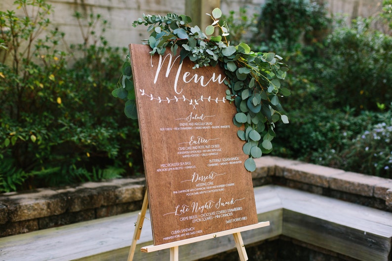 May include: A wooden sign with a white painted menu on an easel. The sign is decorated with greenery. The menu includes the words "Salad", "Entree", "Dessert", and "Late Night Snack".