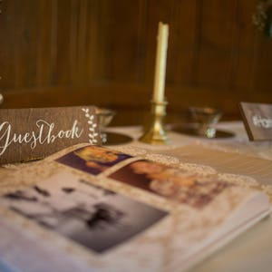 Peut inclure: Une pancarte en bois avec le texte "Guestbook" en &eacute;criture blanche sur fond brun. La pancarte est d&eacute;cor&eacute;e d'accents floraux blancs. Un livre d'or recouvert de dentelle blanche est ouvert sur une table avec des photos de personnes sur les pages.