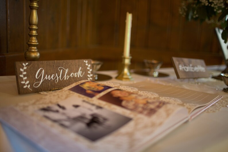 Peut inclure: Une pancarte en bois avec le texte "Guestbook" en &eacute;criture blanche sur fond brun. La pancarte est d&eacute;cor&eacute;e d'accents floraux blancs. Un livre d'or recouvert de dentelle blanche est ouvert sur une table avec des photos de personnes sur les pages.