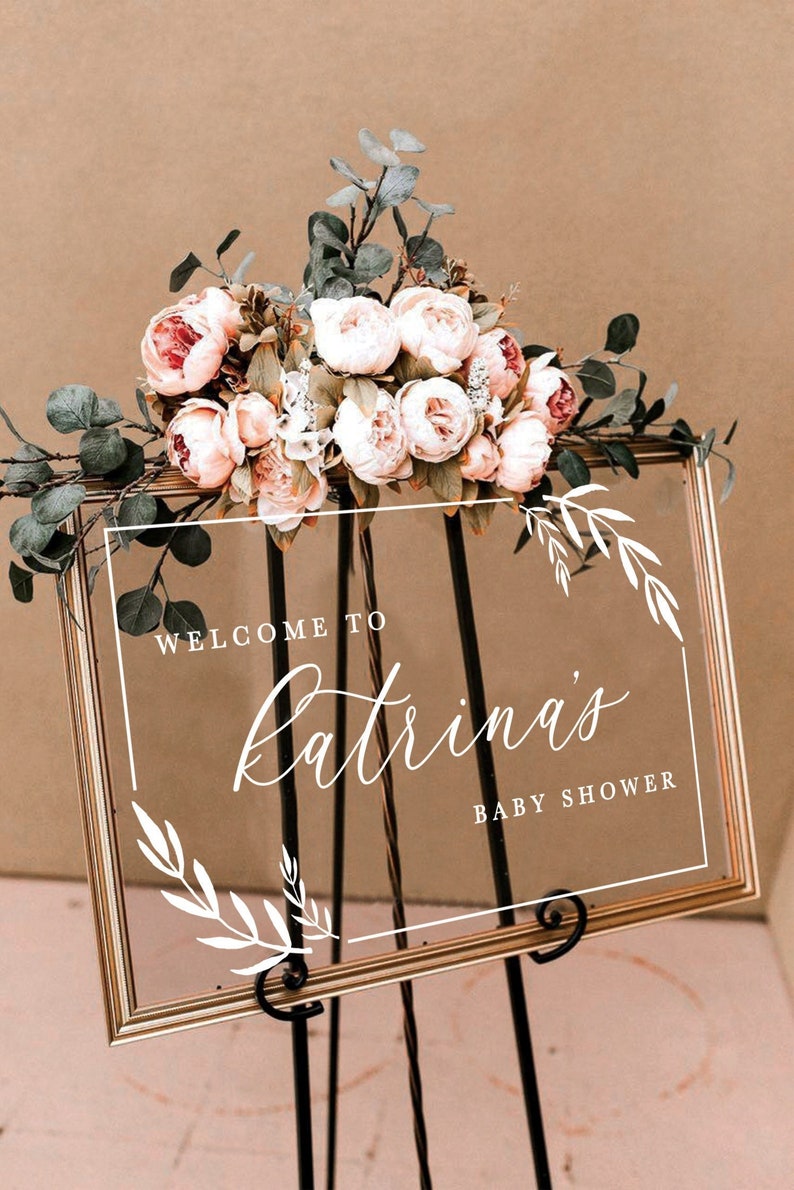 May include: A gold framed clear acrylic sign with white calligraphy that reads "Welcome to Katrina's Baby Shower". The sign is decorated with white floral sprigs and is sitting on a black easel. A bouquet of pink and white flowers with green leaves is sitting on top of the sign.