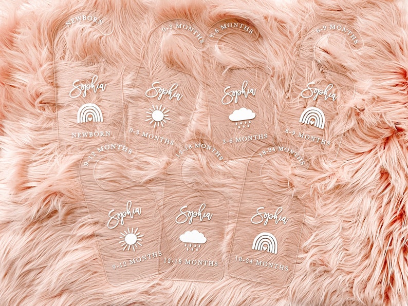 May include: Six clear acrylic baby milestone hangers with the name "Sophia" and different weather icons. The hangers are labeled with age ranges from newborn to 24 months.