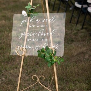 Puede incluir: Un caballete de metal dorado con un letrero de acr&iacute;lico transparente que dice "pick a seat not a side you're loved by both the groom & bride". El letrero est&aacute; decorado con una rosa blanca y hojas verdes.