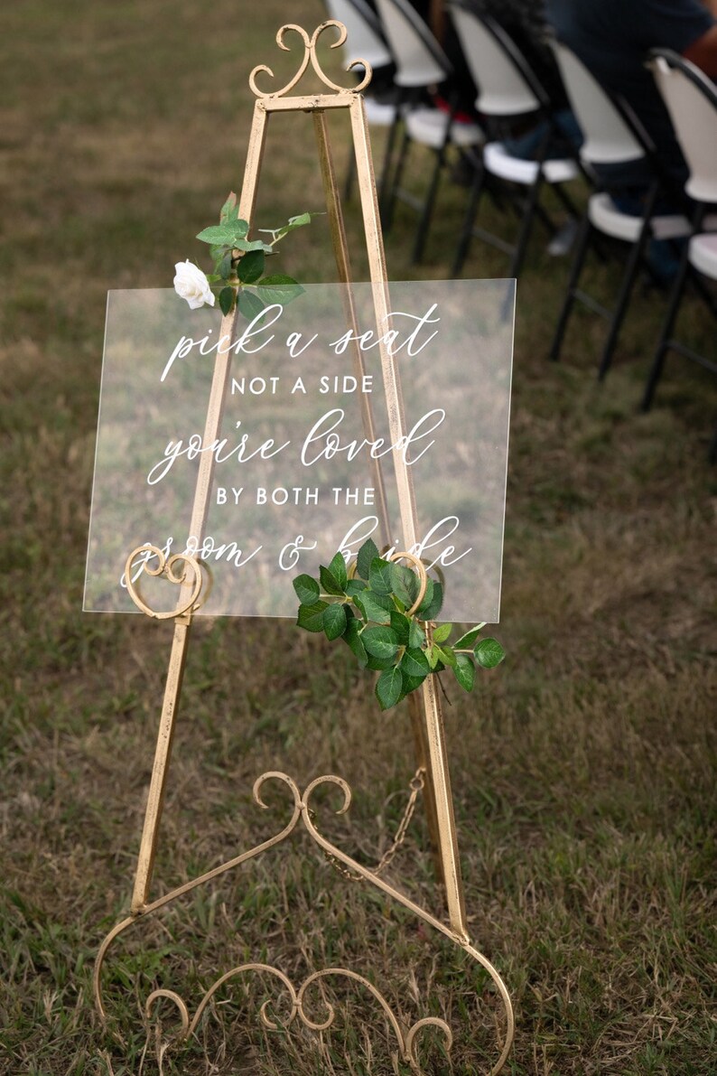 Puede incluir: Un caballete de metal dorado con un letrero de acr&iacute;lico transparente que dice "pick a seat not a side you're loved by both the groom & bride". El letrero est&aacute; decorado con una rosa blanca y hojas verdes.
